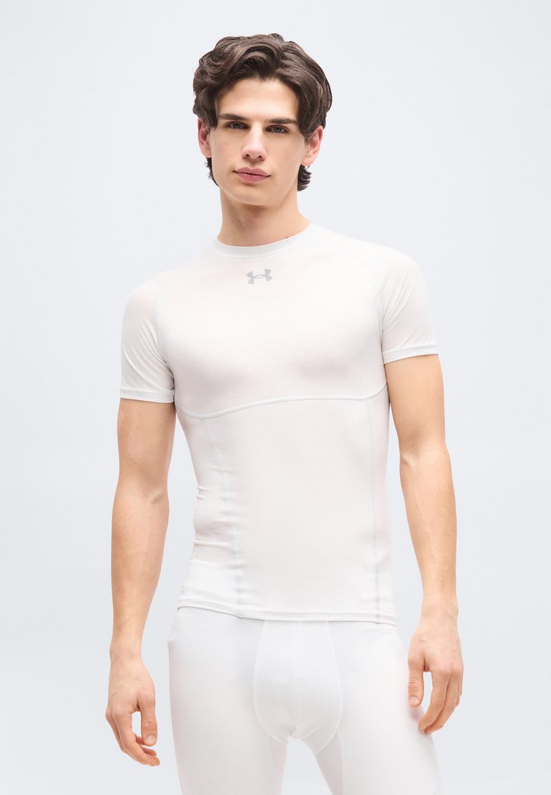 Νεαρός άνδρας με κοντά καστανά μαλλιά, φορώντας στενό λευκό αθλητικό μπλουζάκι και αντίστοιχο περίσφιγκο παντελόνι, στέκεται μπροστά σε απλό φωτεινό φόντο.