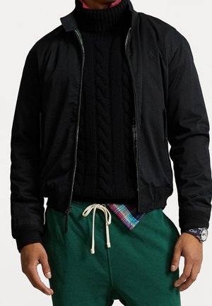 Man draagt een zwarte jas met rits over een zwarte kabelgebreide coltrui, groene broek met aantrekkoord en een polshorloge met een marineblauwe band.