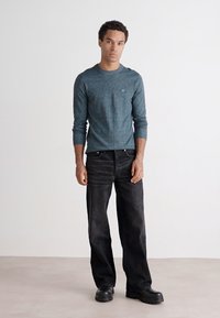Hosszú ujjú, teal színű, texturált anyagú crewneck póló, laza fazonú fekete farmerral és masszív fekete bakancsokkal párosítva.