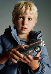 Une voiture jouet en argent tenue par un enfant aux cheveux blonds, portant une veste rembourrée bleue et une chemise rayée, sur un fond neutre.