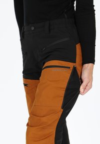 Pantaloni da esterno neri e arancioni con tasche con zip, vita elasticizzata e materiale testurizzato. Presentano un logo e pannelli a contrasto.
