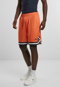Basketballshorts i orange mesh-stof med en sort og hvid stribet kant og logo-detalje på siden; løstsiddende pasform for komfort.