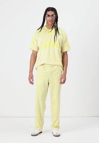 Világos sárga sportöltözet készlet, amely lazán szabott, rövid ujjú pólóból áll, rajta az "adidas" logóval, valamint hozzá illő nadrággal, puha, texturált anyagból.