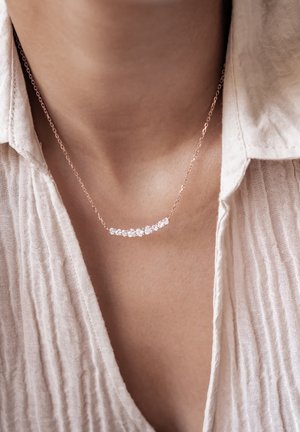 Halskette aus Roségold mit einem geschwungenen Design, verziert mit mehreren klaren Edelsteinen, die auf einem leicht strukturierten beigen Stoff liegt.