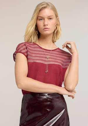 Femme blonde portant un haut rouge à manches courtes avec une partie supérieure rayée transparente, des boucles d'oreilles en or, un collier et une jupe sombre brillante sur fond neutre.