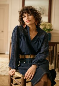 Blouse bleu marine avec manches en dentelle, col en V et détails froncés. Portée avec une jupe en jean et une ceinture beige avec une boucle décorative.