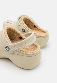 Zuecos de un beige claro con un suave forro de piel, tacón levantado y perforaciones en la parte delantera. Cuenta con una correa para un ajuste seguro y un detalle de logo.