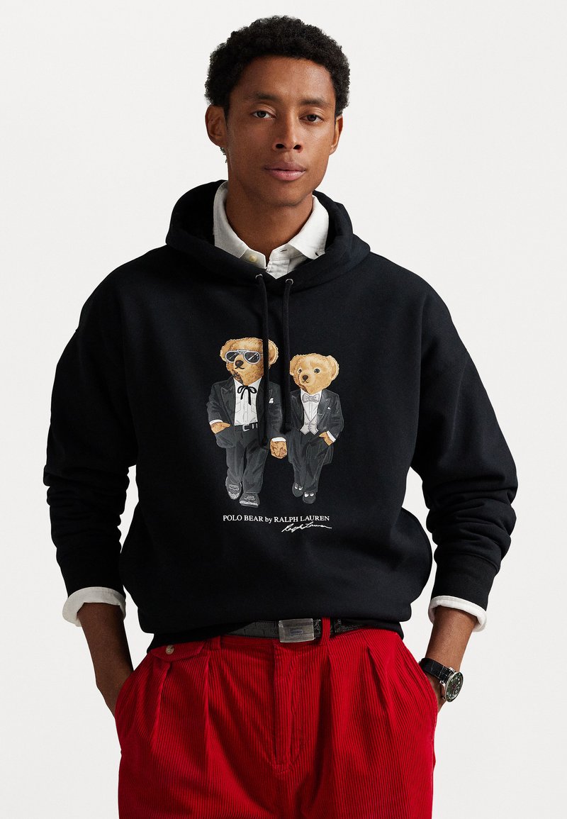 Melns kokvilnas kapucis, uz kura attēloti divi karikatūras lāči tuxedos, viens ar saulesbrillēm, apakšā ir teksts "POLO BEAR by RALPH LAUREN".