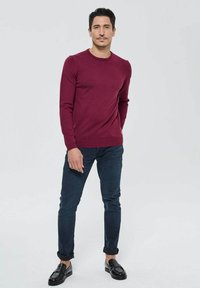 Pull en maille bordeaux avec un col rond, manches longues, associé à un jean foncé et des mocassins noirs. Texture lisse, coupe ajustée.