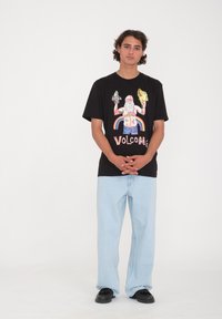 Sort grafisk t-shirt med farverigt design og "VOLCOM" tekst, parret med lys blå brede jeans og sorte sko. Enkel baggrund.