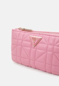 Różowa pikowana skórzana saszetka na zamek z złotym metalowym logo Guess USA i detalami przeszyć na białym tle.