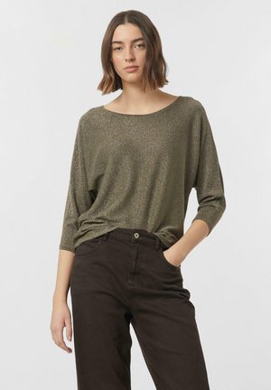 Frau mit schulterlangem braunem Haar, die ein metallisch olivgrünes Langarmshirt und dunkelbraune Hosen trägt, mit der Hand in der Tasche, neutraler Hintergrund.