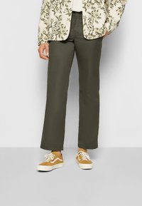 Olive green, tailored pants with a straight leg design, worn with tan Vans sneakers featuring white accents and a floral-patterned blazer.