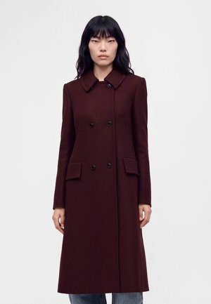 Femme portant un long manteau bordeaux foncé à double boutonnage avec un col et des poches à rabat, debout devant un fond clair uni.