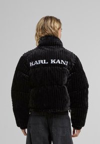 Black, ribbed velvet puffer jacket with a high collar and embroidered "KARL KANI" logo at the back. Features wide sleeves and a cropped design.