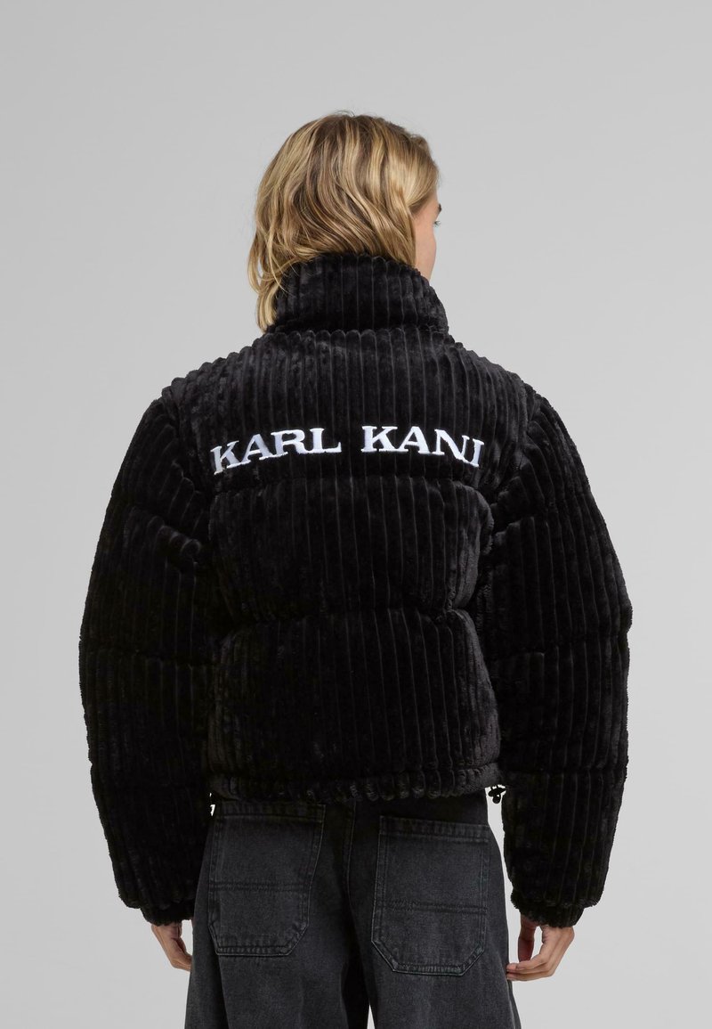 Black, ribbed velvet puffer jacket with a high collar and embroidered "KARL KANI" logo at the back. Features wide sleeves and a cropped design.