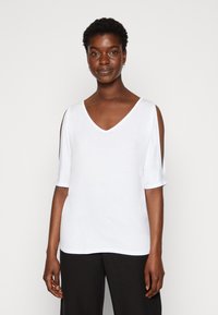 Witte v-hals top met korte mouwen en open schoudernaden, gladde textuur en een ontspannen pasvorm, gecombineerd met zwarte broeken.