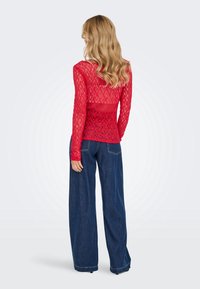 Femme aux cheveux blonds portant un haut rouge en dentelle à manches longues et un jean large bleu foncé, debout de dos sur un fond uni.