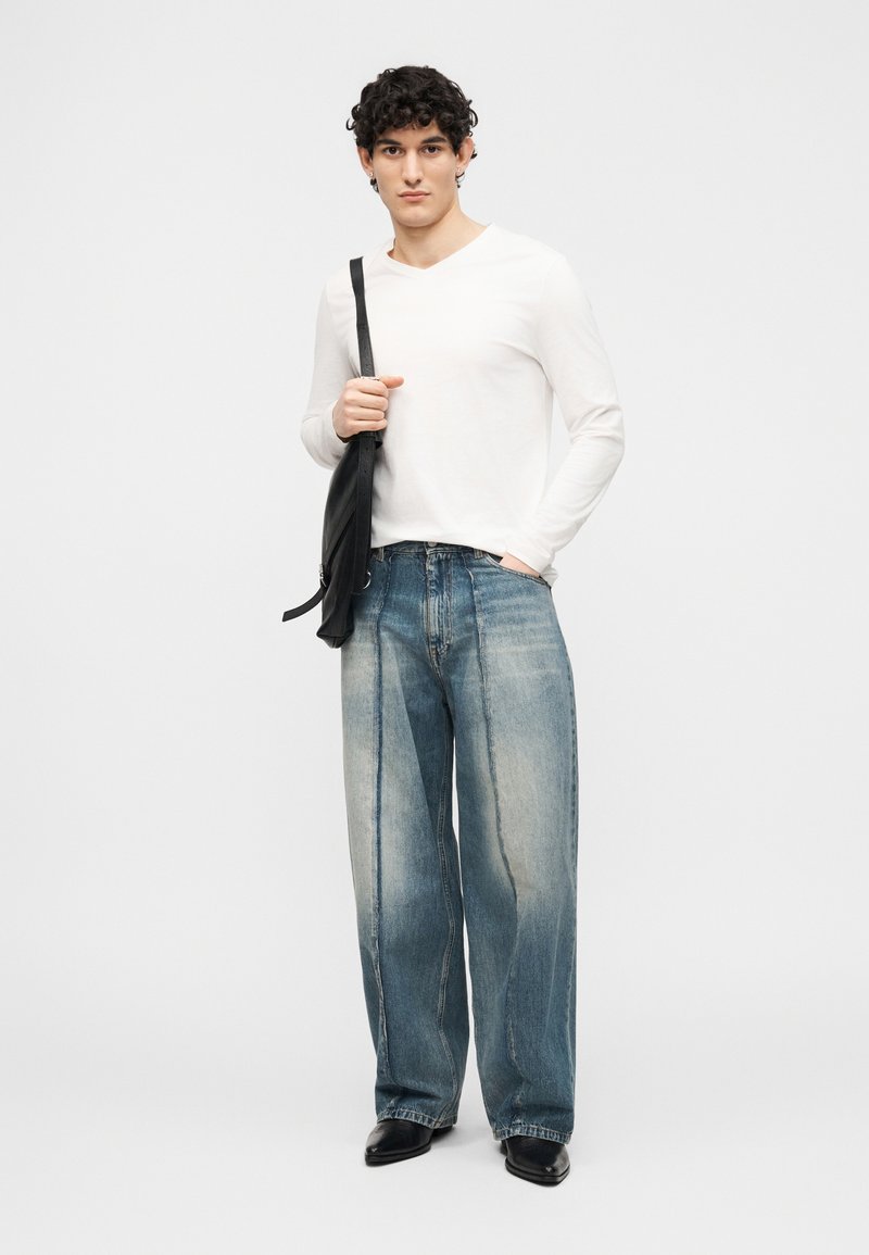 Man met krullend haar, gekleed in een wit shirt met lange mouwen, losse vervaagde blauwe spijkerbroek, zwarte schoenen en met een zwarte schoudertas.