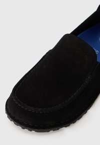 Sorte semskede slip-on sko med rund tå, synlige søm langs kantene og en teksturert gummisåle. Innvendig har den en blå innleggssåle.