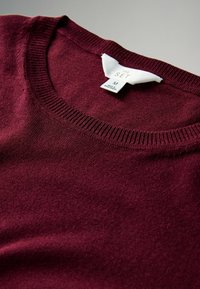 Burgunderfarbener Pullover mit glatter Textur, geripptem Ausschnitt und einem Etikett mit der Aufschrift "THE SET", Größe M, vor einem neutralen Hintergrund präsentiert.