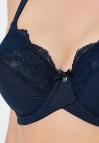 Tummansininen rintaliivi, jossa on pitsikoristelu kuppeissa ja reunassa. Siinä on pieni nauhakoriste ja hopeiset yksityiskohdat keskellä edessä. Pehmeä tekstuuri.