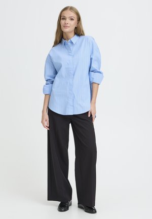 Lichtblauwe, lange mouw blouse met verticale witte strepen, gecombineerd met zwarte brede broek en zwarte schoenen.