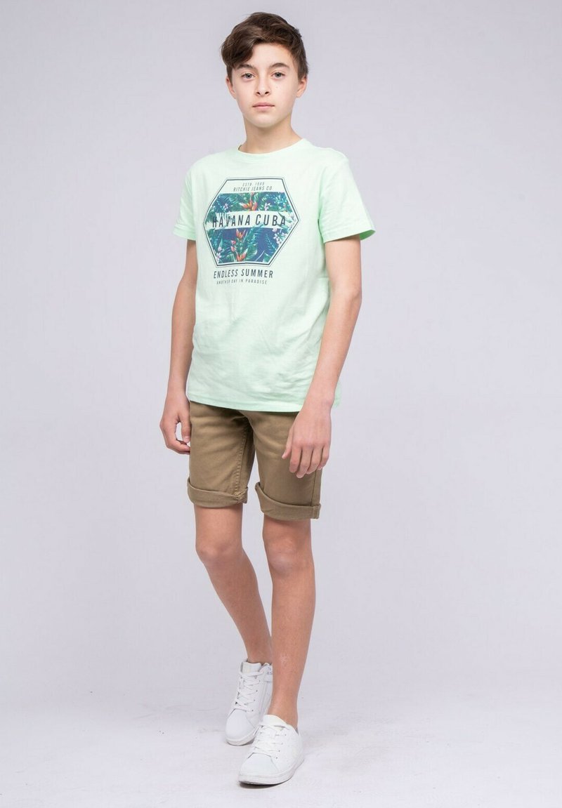 T-shirt vert clair avec impression graphique en forme hexagonale, associé à un short beige retroussé et des baskets blanches. Design simple, style décontracté.