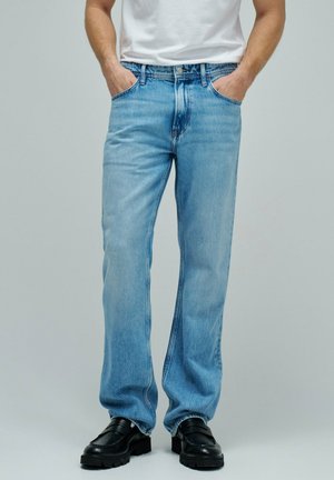 Hombre con vaqueros de pierna recta azul claro, camiseta blanca, manos en los bolsillos y mocasines negros con suela gruesa, sobre un fondo gris liso.