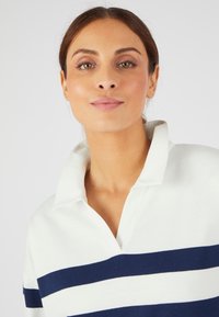 Camisa de polo blanca con rayas horizontales azul marino, con un cuello clásico y una textura de tela suave. No se presentan detalles adicionales.
