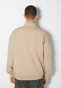 Pullover beige con colletto alto, taglio oversize e polsini a costine. Tessuto morbido, dettagli di cucitura minimi e nessun logo visibile.