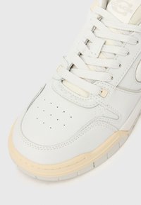 Fehér bőr sportcipő közeli képe, lyukacsos díszítéssel, vastag fehér fűzőkkel és bézs gumitalp szegéllyel, semleges háttéren.