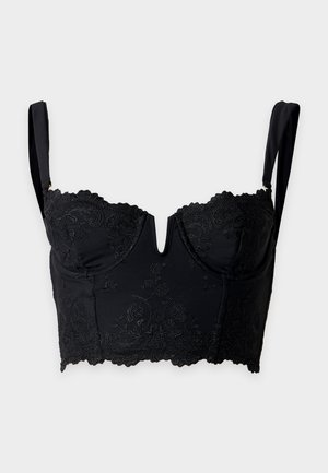 Femme debout face à l’avant, portant un crop top en dentelle noire, des sous-vêtements assortis et des tongs plateformes noires sur un fond clair uni.