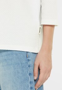 White textured top with three-quarter sleeves, paired with light blue jeans. Tag on the side reads "SENSES."