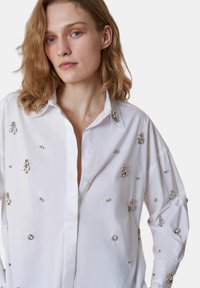 Camicia bianca con colletto decorata con embellissements in argento, caratterizzata da una vestibilità ampia e maniche lunghe, realizzata in un tessuto leggero e liscio.