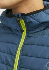 Giacca a vento blu con finitura texturizzata, dotata di un colletto nero e una cerniera verde brillante. Include un dettaglio logo ben visibile.