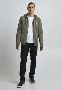 Hoodie zippé vert olive en tissu texturé, avec des poignets et un ourlet côtelés ; porté par-dessus un t-shirt blanc, associé à un pantalon noir et des baskets.