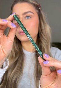 Groene eyelinerpotlood met een gladde afwerking, voorzien van een ronde punt en merktext op het onderste deel. Nagels zijn gelakt in lavendel.