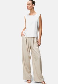 White sleeveless top with textured neckline, paired with loose beige pleated trousers. Model stands with one hand in pocket, wearing light sandals.
