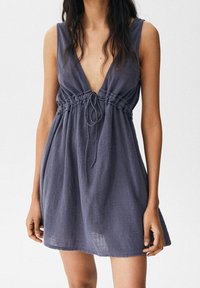 Robe sans manches à décolleté en V plongeant, de couleur gris foncé, en tissu léger et texturé. Présente une taille froncée avec un cordon de serrage pour un ajustement.