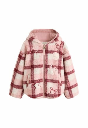 Mädchen mit lockigem Haar, das eine rosa-rote karierte Reißverschlussjacke, weite blaue Jeans und weiße Turnschuhe trägt, steht auf einem grauen Boden.