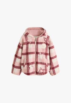 Mädchen mit lockigem Haar, das eine rosa-rote karierte Reißverschlussjacke, weite blaue Jeans und weiße Turnschuhe trägt, steht auf einem grauen Boden.