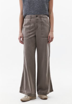 Osoba nosící široké kalhoty barvy taupe s předními náprsními kapsami, spojené s béžovými teniskami a šedým tílkem na bílém pozadí.