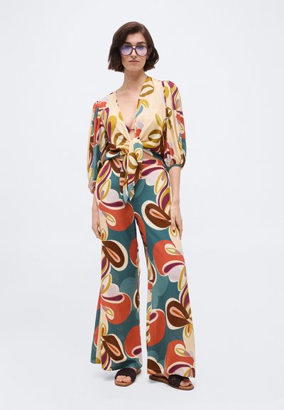 Γυναίκα που φοράει ταιριαστό πολύχρωμο μπλουζάκι με floral σχέδιο, ζωνάκι μπροστά και παντελόνια με φαρδιά πόδια, σκούρα γυαλιά ηλίου και μαύρα σανδάλια.