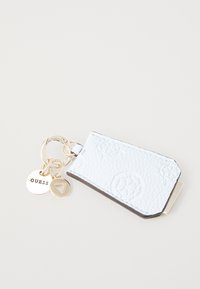 Lys blå faux skinnnøkkelholder med preget design, hardware i gulltone og festede merkelapper, inkludert et rundt "GUESS" emblem.