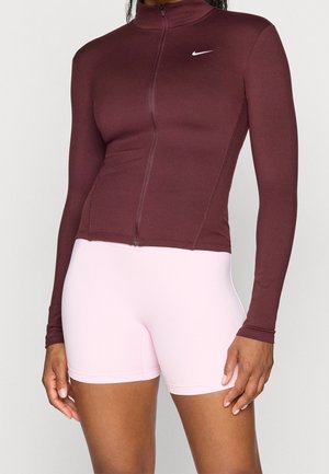 Femme portant un haut zippé ajusté bordeaux à manches longues avec un logo Nike blanc et un short moulant rose clair.