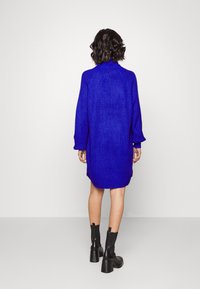 Oversized blå stickad tröjdress med hög krage och långa ärmar, med ribbad mudd och något rundad fåll. Svarta chunky ankälskor.