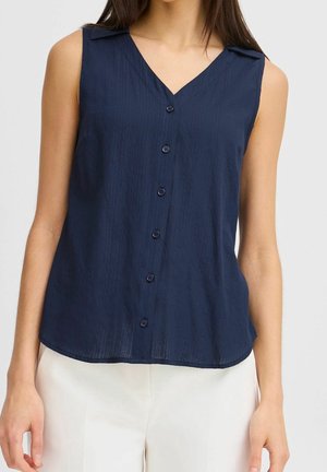 Blouse bleu marine sans manches avec boutons et col en V portée avec un pantalon blanc sur une personne debout.