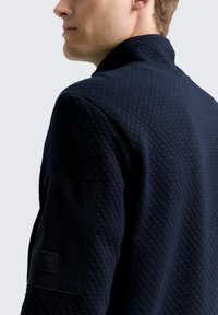Chaqueta texturizada azul marino con un cuello alto y un bolsillo tipo parche en la manga izquierda. Presenta un patrón acolchado y acentos de tela suave.
