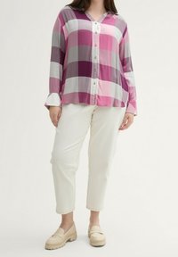 Kvinde iført en lyserød og lilla ternet skjorte med knapper, hvide cropped bukser og beige loafers, stående mod en simpel lys baggrund.